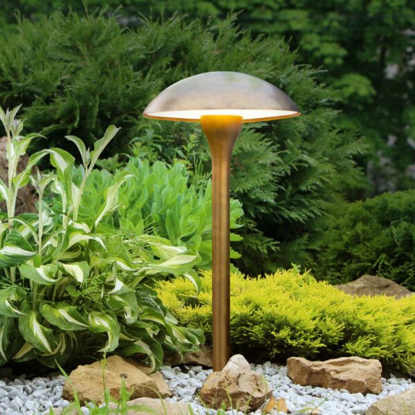 Image of weathered brass Fungy garden light in daytime, shown on rockery with planting in the background