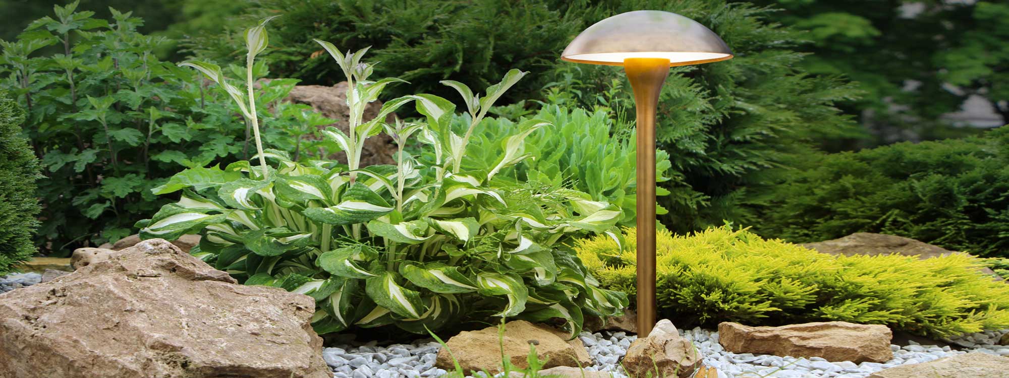 Image of Royal Botania Fungy bollard downlight in weathered brass, shown on rockery with planting in the background