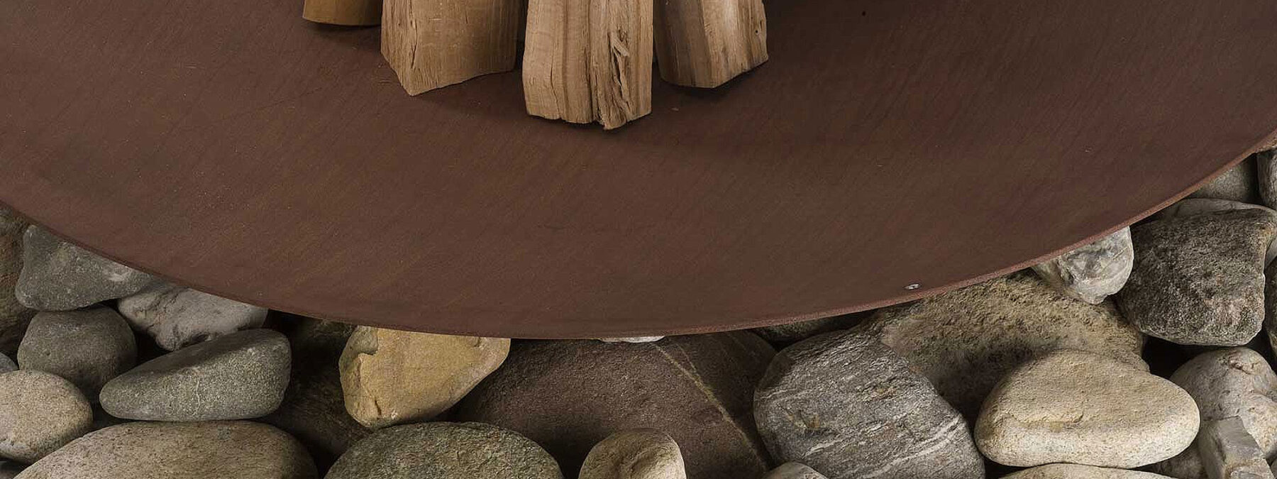 Image of Discolo corten fire bowl by AK47 stacked with logs sat upon large stones