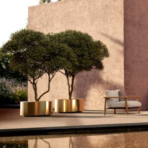 Image of late afternoon sun glinting on Cuprum large brass planters, shown next to Mambo teak lounge chair and tranquil water feature in the foreground