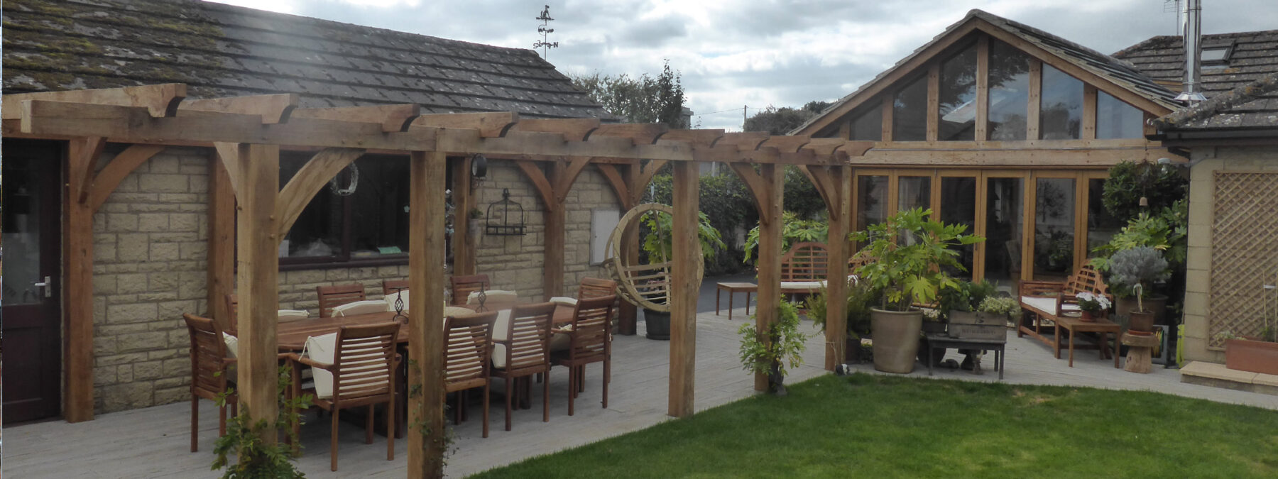Image of Oakcraft luxury oak pergola with dining furniture beneath and a swing seat in the background