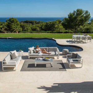 Image of woman lying on Reno modern 3 seat garden sofa by Jati & Kebon, with swimming pool, woodland and sea in the distance beneath blue sky