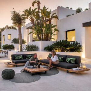 Image of Truro Lounge FSC teak corner sofa on minimalist terrace at dusk, with palm trees and white-washed building in background